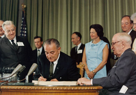 President Lyndon B. Johnson signing the Medicare bill at the Harry S. Truman Library in Independence Missouri. Former President Harry S. Truman is seated at the table with Johnson. In the background are Sen. Edward V. Long an uniden