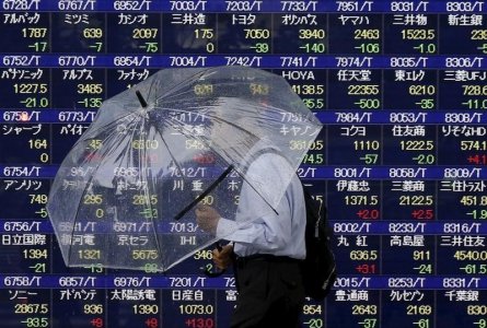 A man holding an umbrella walks in front of an electronic stock quotation board outside a brokerage in Tokyo