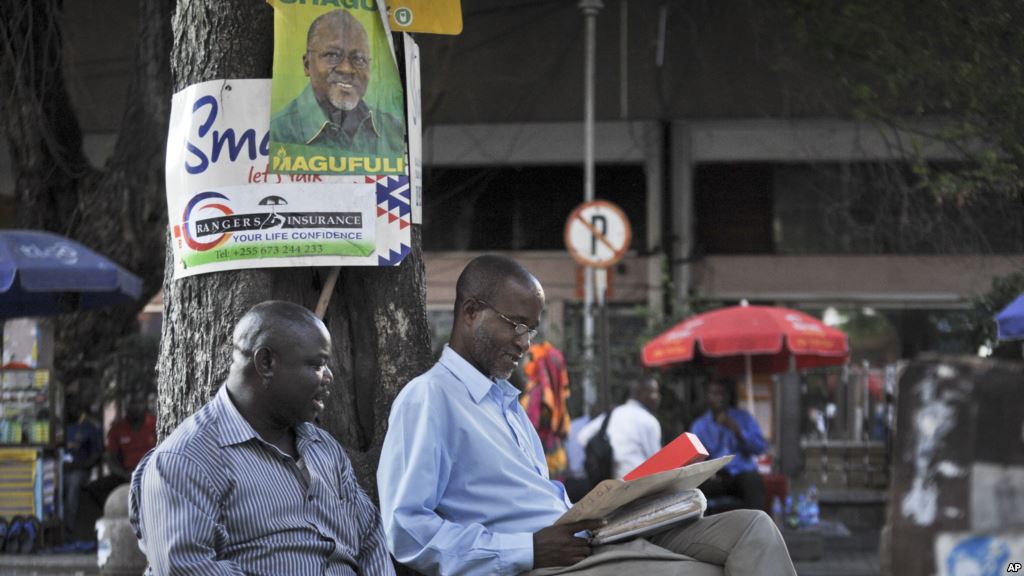 Tanzanians sit next to a tree underneath an election poster for ruling party presidential candidate John Magufuli as they await election results in Dar es Salaam Tanzania Oct. 27 2015