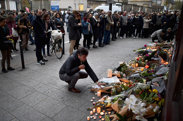 A man kneels in front of candles and flowers as mourners gather in front of the Petit Cambodge and Le Carillon restaurants