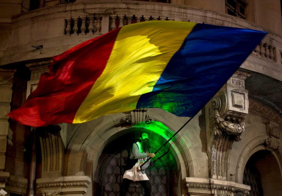 A man's face is illuminated by a laser torch as he waves a large Romanian flag after climbing the University building during the third day of protests joined by tens of thousands across the country calling for early elections in Bucharest Romania T