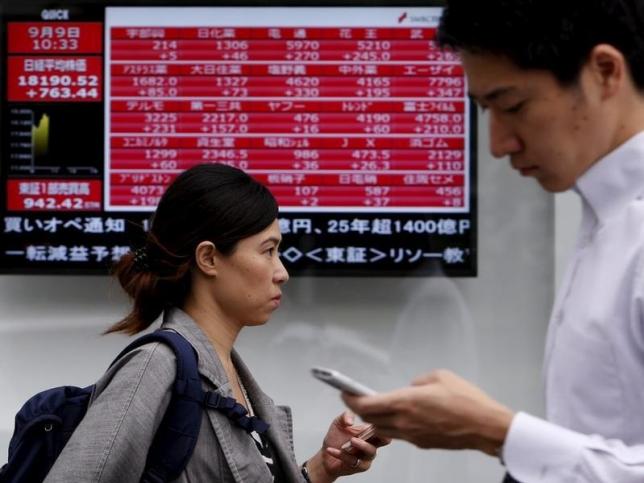 Pedestrians holding their mobile phone walk past an electronic board showing the various stock prices outside a brokerage in Tokyo Japan