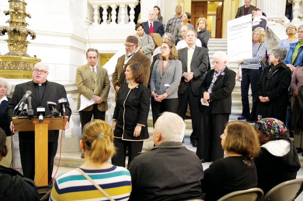 COMMON GROUND Father Bernard Healey addressed the crowd at a State House rally in favor of Syrian refugee resettlement last Thursday as immigrants resettled by the diocesan Office of Immigration and Refugee Services look on from the front row. Other fait