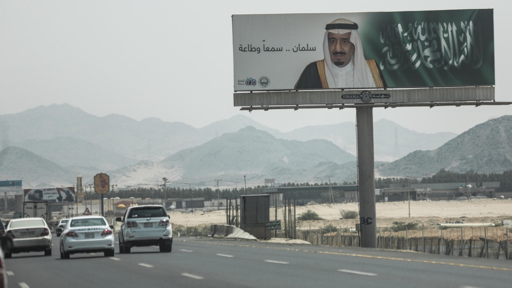 Cars drive past a banner of Saudi Arabia's King Salman that reads'Salman we hear and obey' near Mecca on Sept. 17. A drop in oil revenue is forcing Saudi Arabia to consider cuts to social programs. The Saudis have declined to cut production a