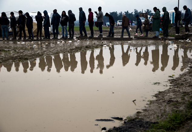 Migrants are reflected in a puddle while waiting in line to receive hot tea during a rainfall at the northern Greek border point of Idomeni Greece Tuesday