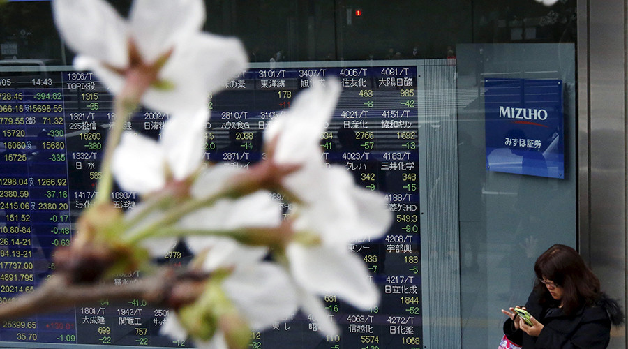 A woman uses a smart phone next to an electronic stock quotation board as cherry blossoms bloom outside a brokerage in Tokyo Japan