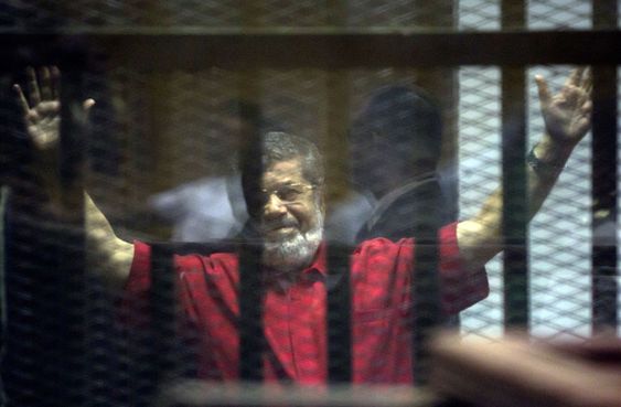 Mohammed Morsi wearing a red jumpsuit that designates he has been sentenced to death raises his hands inside a defendants cage in a makeshift courtroom at the national police academy in an eastern suburb of Cairo Egypt