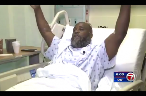 Black man in a white hospital gown sits in a bed with his hands above his head