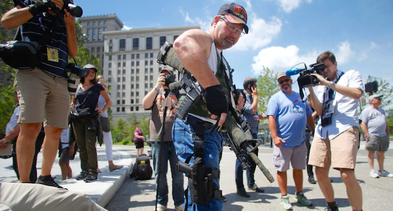 Steve Thacker carrying a rifle and a handgun is surrounded by news media in a public square in Cleveland