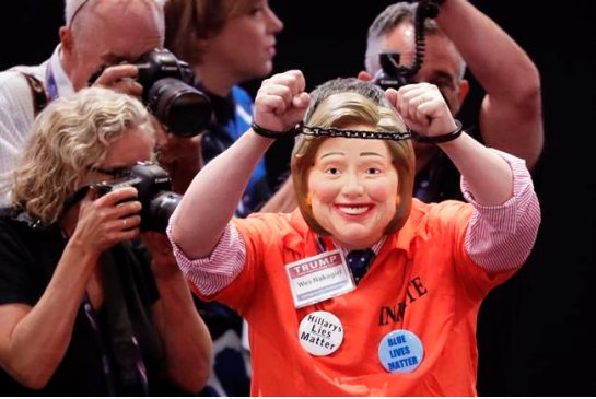 Wes Nakagiri wears a Hillary Clinton mask and holds up handcuffs as he walks across the floor before the start of the final session of the Republican National Convention in Cleveland Thursday