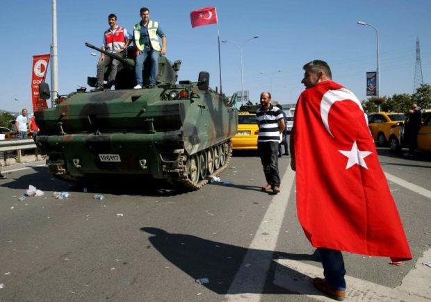 A man wrapped in a Turkish flag walks past a military vehicle in front of Sabiha Airport in Istanbul