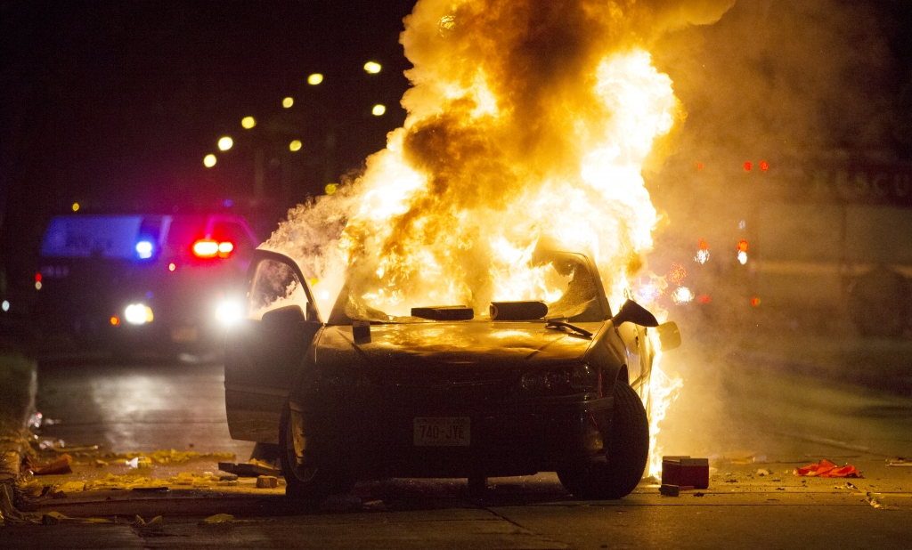 A car burns as a crowd of more than 100 people gathers following the fatal shooting of a man