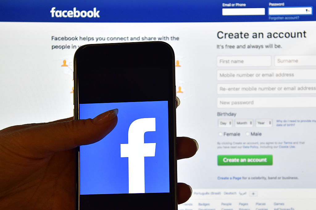 LONDON ENGLAND- AUGUST 03 A person holds an iPhone displaying the Facebook app logo in front of a computer screen showing the facebook login page