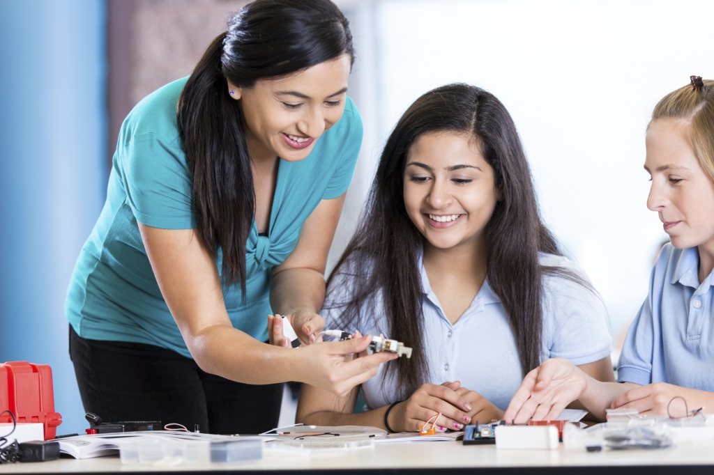 Mid adult Hispanic woman is teaching middle school or junior high school class about audio engineering. Teacher is using electronic or robotic parts to help students create speakers. Preteen Indian and Caucasian girls are smiling while studying STEM. Scie