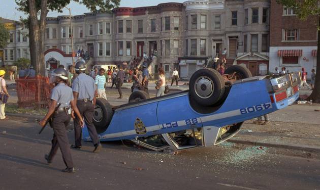 New York Police Department officers in riot gear walk past a police car that was overturned by rioters in the Crown Heights section of the Brooklyn borough of New York. A plan to mark the 25th anniversary of the