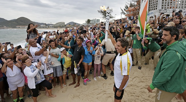 Brazilian Rico de Souza celebrates with the crowd as he prepares to surf with the Olympic torch at Macumba beach as the torch relay continues on its journey to the opening ceremony of Rio's 2016 Summer Olympics in Rio de Janeiro Brazil Thursday A