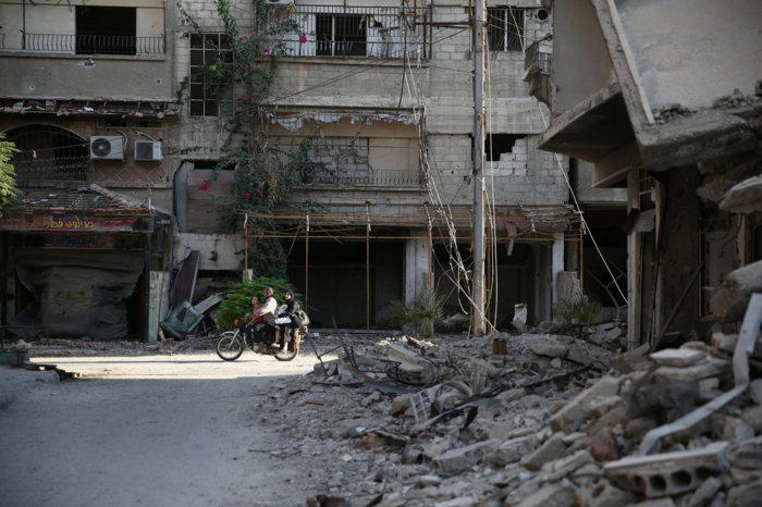 People ride a motorcycle near damaged buildings in rebel held Ain Tarma