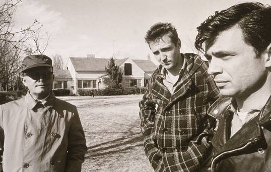 American novelist Truman Capote, dressed in an overcoat glasses and hat stands on a lawn near American actors Scott Wilson and Robert Blake, both dressed in character while on location filming In Cold Blood