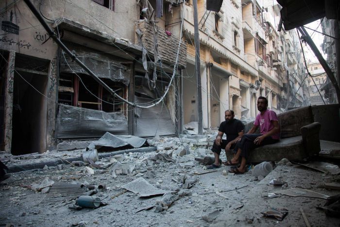 Two men sit on an old couch looking at the rubble around them