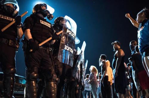 Police in black riot gear near protesters against dark night sky